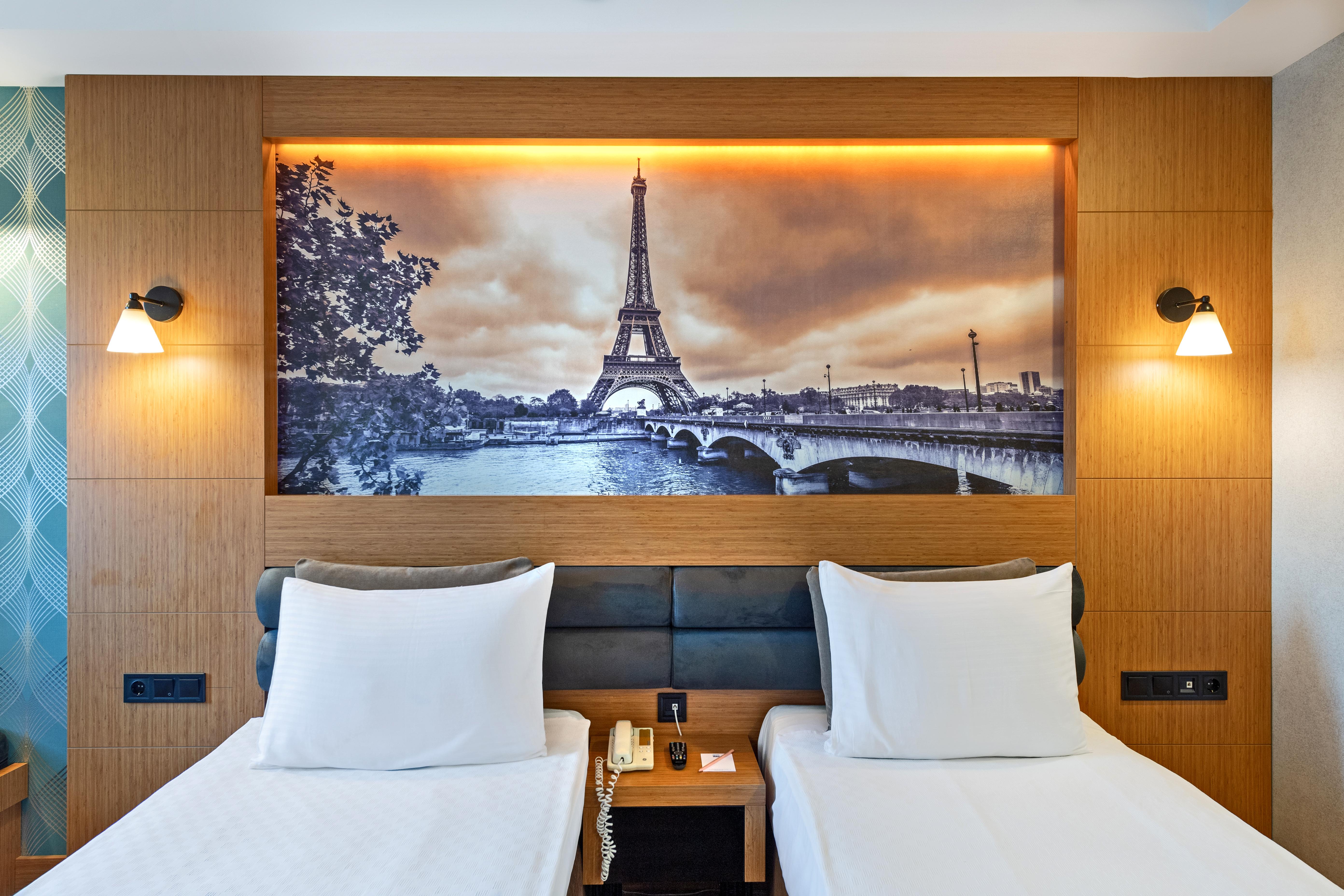 Sealife Family Resort Hotel Antalya Eksteriør billede The photo depicts a hotel room featuring two single beds with white bedding. Behind the beds, there's a large mural of the Eiffel Tower, set against a scenic backdrop of the Seine River. The image has a vintage, sepia-toned appearance, enhancing the 