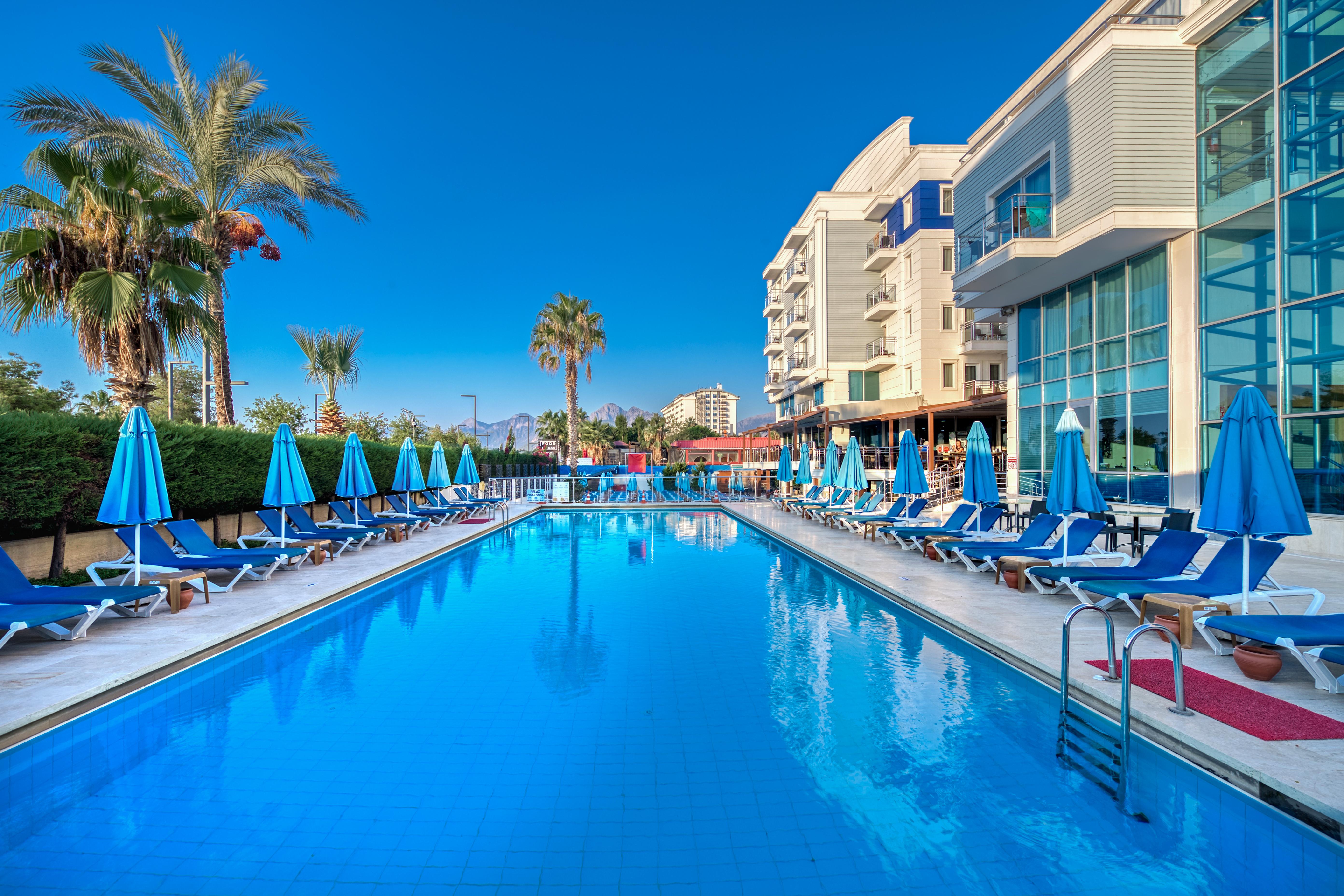 Sealife Family Resort Hotel Antalya Eksteriør billede The photo depicts a serene poolside scene at a hotel or resort. In the foreground, a large blue swimming pool reflects the clear sky, with comfortable lounge chairs arranged neatly around it under blue umbrellas. Lush palm trees add to the tropical a